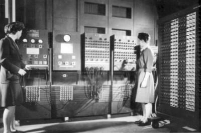 Au fond de la pièce, une grosse machine, l’ordinateur ENIAC. La machine est composée de 7 panneaux, recouverts de boutons, d’indicaterus lumineux et de câbles. On peut voir un amas de câbles branchés sur l’ordinateur. Devant la machine se tiennent debout deux femmes, Betty Jenning à gauche et Frances Bilas à droite. Frances Bilas règle un bouton rotatif, tandis que Betty Jenning semble bouger des cables.