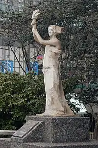 Photo couleur d'une statue beige représentant une femme debout, bras nus, chevelure au vent, et portant, de ses deux mains, une torche.