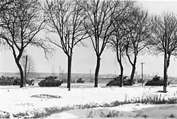 Photo noir et blanc d'une colonne de chars dans la neige, derrière des arbres.