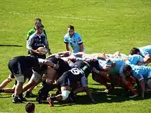 Des joueurs de rugby engagent une mêlée, sous le regard de l'arbitre et des demis de mêlée.