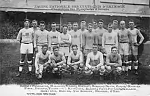 Photographie noire et blanche d'une équipe de rugby devant une tribune. Les joueurs sont répartis sur deux rangées.