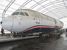 Photo de l'avant du corps de l'avion, dans un grand hangar, avec des traces de boues et d'impact d'oiseaux visibles, notamment avec un trou sur son nez.