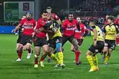 Photographie d'un joueur avec le ballon dans les main, lors d'un match de rugby entre une équipe rouge et une jaune.