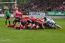 Mêlée. Les rouges (Oyonnax) poussent à gauche. Les ciel et blanc poussent à droite, Lauret en premier plan, à sa place de flanker. Le demi de mêlée rouge et noir attend le ballon. L'arbitre et le demi de mêlée ciel et blanc (Machenaud) observent.