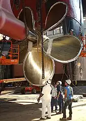 Vue du chantier naval, zoom sur une hélice de propulsion. Les hommes en dessous donnent une idée de la taille de l'hélice.