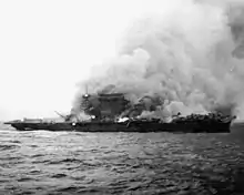 D'énormes colonnes de fumée s'élèvent d'un porte-avions fortement incliné sur tribord. Plusieurs foyers d'incendie sont clairement visibles sur l'ensemble du navire.