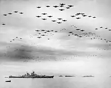 Photographie en noir et blanc montrant un grand nombre d'appareils volant en formation au-dessus de navires de guerre