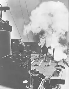 Photographie de l'avant du navire prise depuis le château. La tourelle la plus en avant dont les canons sont relevés vient de tirer.