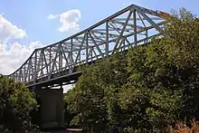 Vue d'un pont métallique enjambant un cours d'eau. Des arbres sont présents de part et d'autre.