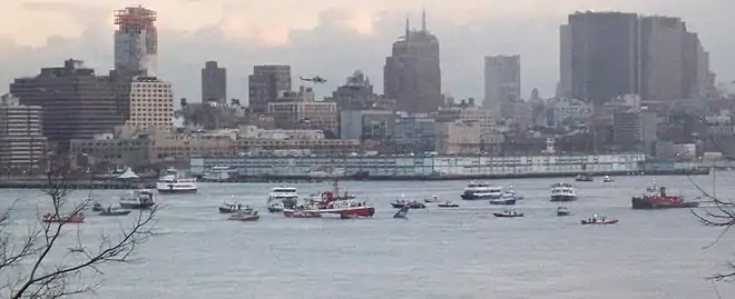 Photo montrant de nombreux navires au milieu du fleuve Hudson, près de l'avion, situé au centre de l'image, et dont seule sa queue est visible, le reste se trouvant sous l'eau. En arrière-plan se trouve la ville de New York.