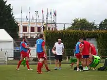 Des joueurs de rugby en chasubles avec du matériel d'entraînement sur un terrain ; en arrière-plan, des arènes décorées avec des drapeaux.