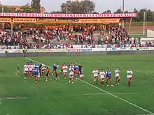 Des joueurs de rugby marchent sur la pelouse ; en arrière-plan, des supporteurs applaudissent depuis une tribune.