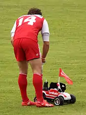 Un joueur de rugby se penche pour récupérer un tee et une bouteille d'eau transportés par une voiture télécommandée équipée d'une peluche à l'effigie d'un taureau.