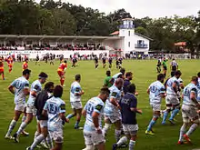 Des joueurs s'entraînent sur le bord d'un terrain alors qu'un match de rugby s'y déroule.