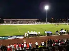 Un match de rugby joué de nuit, sur un stade éclairé. Les spectateurs dans la tribune et autour du terrain sont tous habillés en blanc et rouge.