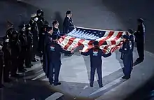 Photographie d'un drapeau américain déchiré tenu par huit personnes devant des militaires, dans un stade la nuit.