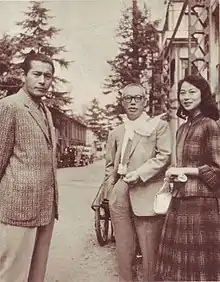 Photographie en noir et blanc de trois individus japonais, qui regardent l’objectif avec le sourire, deux hommes et une femme. Les deux hommes sont en costumes. La scène a lieu dans une rue, le jour.
