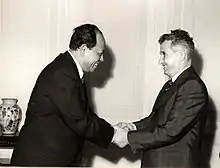 Ieng Sary à gauche serre les mains de Nicolae Ceaușescu à droite; à l’extrême gauche un vase trône sur un guéridon