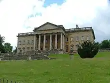 Beau manoir en haut d'une pente gazonnée, avec porche à colonnes ; quelques arbustes verts ; vue légèrement de gauche