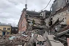 Photo d'un immeuble en briques de trois étages à moitié effondré, deux personnes en uniforme se tenant dans la partie détruite, une partie d'un engin de levage