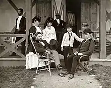 Photographie de famille prise sur le perron d'une maison en bois. Grant en costume et avec un haut-de-forme à la main est assis dans une chaise en osier face à un jeune homme assis dans une chaise pliable en bois et portant un chapeau melon. Derrière eux se trouvent deux femmes en robe et deux garçons.