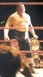 Catcheur torse nu avec une peinture faciale tenant une ceinture de champion dans une main.