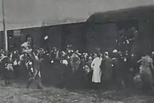 De nombreuses personnes montent dans des wagons. Un policier juif, avec sa casquette et son brassard, marche le long de la foule.