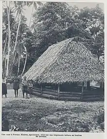 Photographie en noir et blanc montrant une habitation délimitée par des rondins de bois, avec un toit très pentu réalisé en pandanus. Trois femmes et un enfant se tiennent debout à gauche de la maison, on voit des cocotiers et d'autres arbres en arrière-plan.