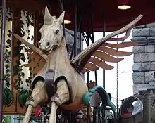 Vue de face d'un cheval mécanique en bois articulé, avec une corne et des ailes.