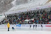 Un juge habillé en jaune se tient à côté d'une chaise. En arrière-plan, des patineurs concourent.