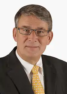 Photographie d'un homme aux cheveux gris, portant des lunettes, un veston et une cravate jaune.