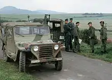 Un groupe de trois soldats américains dont l'un porte un fusil, et deux officiers ouest-allemands de la Bundesgrenzschutz, se tiennent debout à côté de deux véhicules garés sur une étroite bande d'asphalte, devant un paysage composé de champs et de collines.