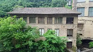 Vue de face de l'usine d'Entraygues sur 3 niveaux avec ses 7 grandes fenêtres carrées ou rectangulaires par étage et son toit en tuiles sombres, beaux arbres au premier plan