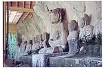 Photographie d'une rangée d'environ. dix statues de pierre assise en face d'un rocher. Vue de trois-quarts. L'une des statues est environ deux fois plus grande que les autres.