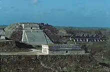 Photographie représentant sur fond de ciel bleu une pyramide de style maya derrière un temple rectangulaire et plat, le tout en pierre grise