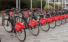 Photographie en couleur représentant une dizaine de vélo noir et rouge accrochés à leur borne de stationnement