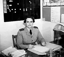 Photographie en noir et blanc représentant une femme vêtue d'un costume militaire, assise dans un bureau.