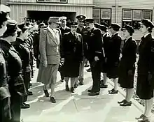 Deux rangées de femmes en uniformes militaires sombres se faisant face, avec un certain nombre d'autres militaires et un civil marchant entre elles.