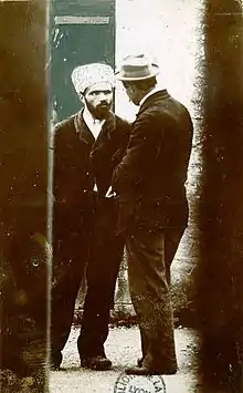 Carte postale couleur ancienne montrant deux hommes debout, se faisant face (tenues sombres). Celui de dos porte un chapeau (un panama) et un paire de moustaches ; celui de face, une barbe et un bonnet (un papakha) de couleur claire.