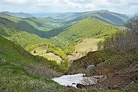 Photo en couleurs d'une vallée entourée de moyennes montagnes très verdoyantes avec des ombres de nuages et un peu de neige au premier plan