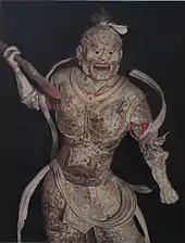 Statue féroce, corps cambré, bras droit levé tenant une lance. Couleur un peu effacée à dominante rouge et or.