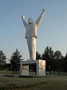 Dans un parc, une statue en pierre blanche représentant la silhouette stylisée d'un homme en uniforme, qui lève ses deux bras en signe de défi.