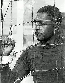 Photo en noir et blanc d'un Afro-Brésilien portant un maillot à manches longues et regardant latéralement à travers une grille.