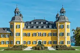 Une manière de château à la belle façade jaune et des tourelles aux angles, un jardin, sans doute à la française, devant, et on aperçoit un morceau de lac derrière.