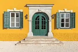 Une façade jaune tirant sur le orange ; une porte d'entrée, au dessus d'un petit perron de 3 marches, qui semblent être en marbre ; de chaque coté, des fenêtres ; chaque percée dans le mur est encadrée d'une sorte de la même matière que les marches.