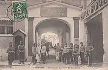 Carte postale avec une photo noir et blanc montrant une entrée gardée par une douzaine de soldats portant dolman à brandebourgs.