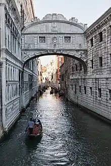 Photo d'un canal bordé de palais vénitiens que traverse une gondole, et surplombé par un pont couvert.