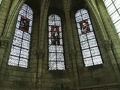Trois vitraux vus de loin, représentant deux hommes et une femme, tous auréolés. Entre eux, des fenêtres en verre blanc et des piliers de pierre.