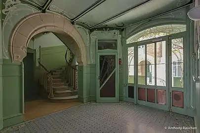 Photo en couleur d'un espace dans les tons verts ouvrant à droite sur une cour et à gauche, sous un arc en pierre, sur un escalier