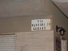 Photographie d'un angle de rue en plan rapproché avec peint sur le mur un rectangle blanc, avec inscrit dessus en lettres majuscules noires : Via Martiri di Gerace.